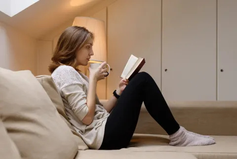Kvinde drikker af et krus og læser en bog på sofaen.