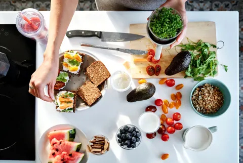 Overhead af kvinde, der laver mad og forbereder sundt måltid med frisk avocado, grøntsager, vandmelon, blåbær og anden frugt i køkkenet