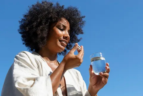   Sort kvinde tager to multivitaminkapsler og holder et glas vand med en blå himmel i baggrunden
