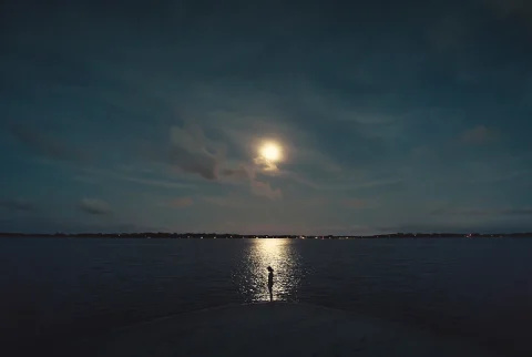   Ung kvindelig silhuet ved kysten med det reflekterede lys fra månen på havet